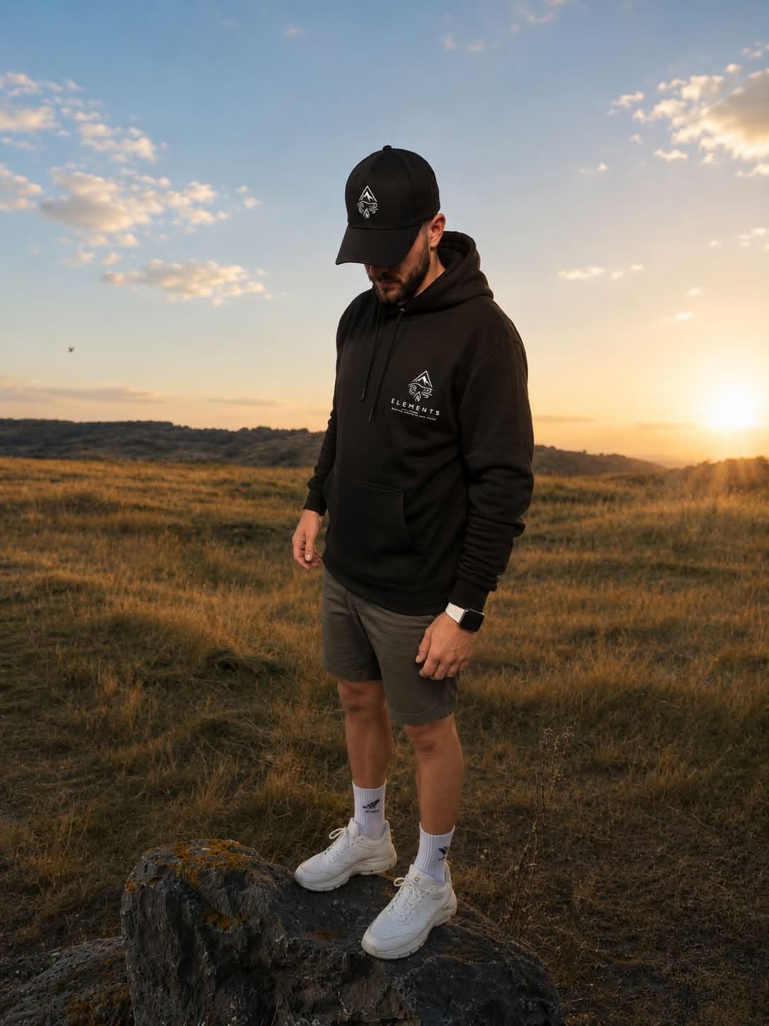 Person in a black Elements hoodie and cap standing on open moorland at sunset