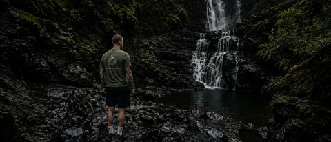Person seen from behind wearing an olive green Elements T-shirt near a waterfall, wide crop for mobile