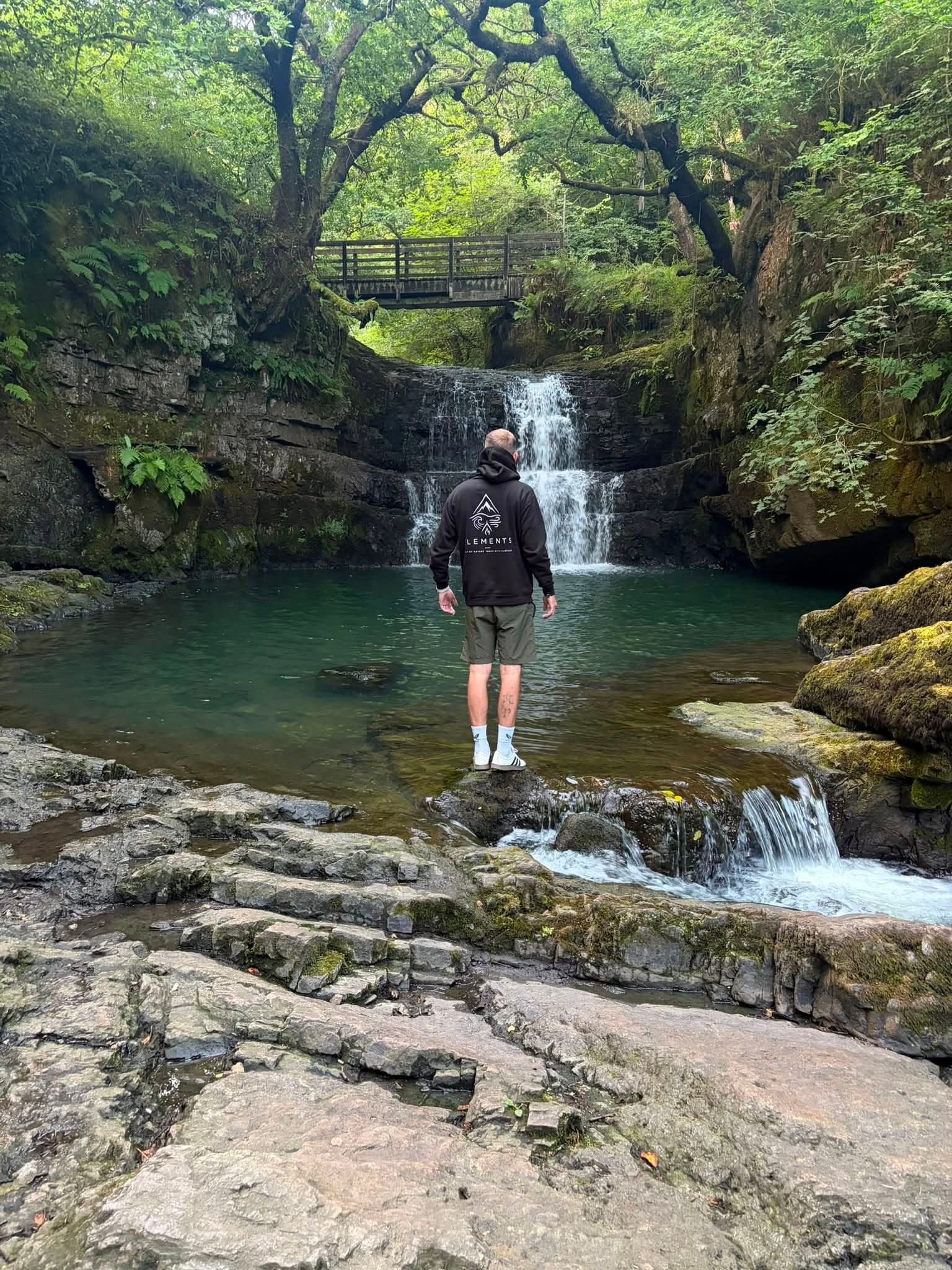 Person in dark clothing and a cap, carrying a rucksack, standing on rocks at the edge of a waterfall pool in a green wooded gorge