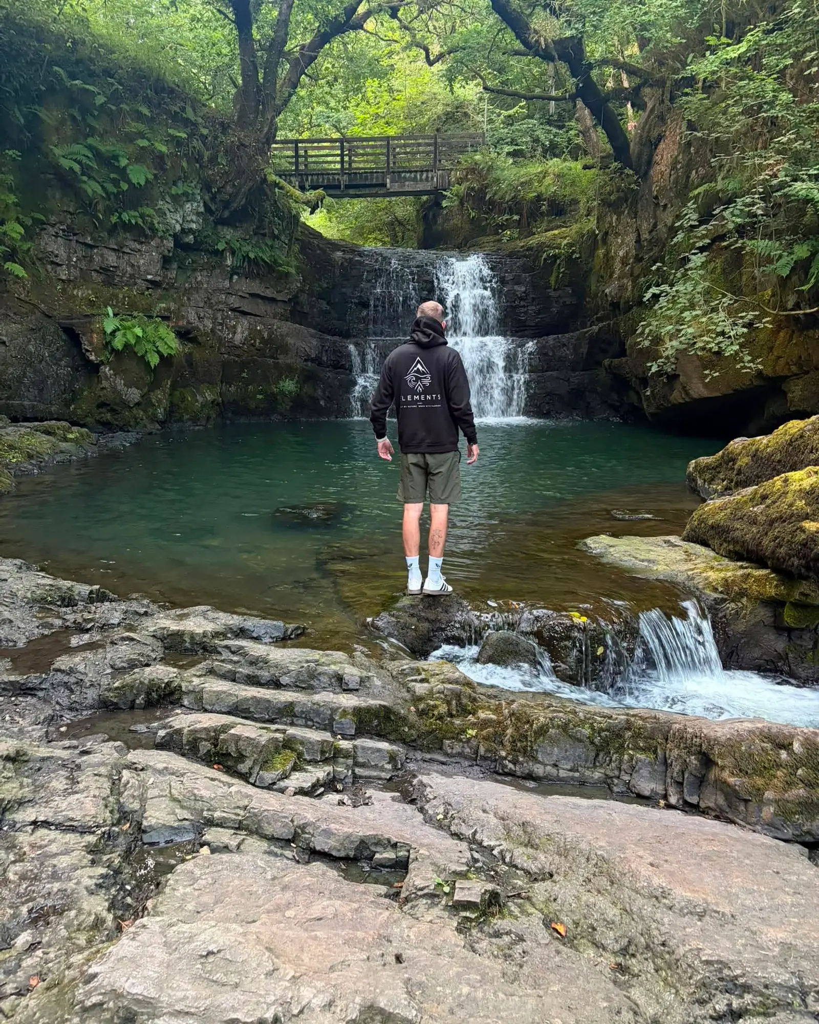 A hiker in an Elements soft-shell, standing before a waterfall in a wooded gorge.